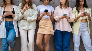 Teknoloji kullanan Z jenerasyonu insanları. Duvara yaslanmış akıllı telefon ekranlarından sosyal medya içeriklerine bakan kızlar. Orta bölüm mahsulü.