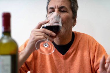 Olgun bir adam beyaz duvara karşı tek başına kırmızı şarap içiyor. Önde bulanık bir şişe var.