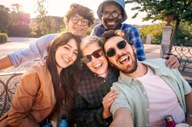 Bir grup mutlu genç arkadaş halk parkında gülerek ve selfie çekerek sıcak bir altın saat gün batımında.