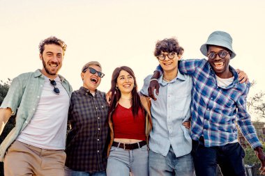 A cheerful group of young multiethnic friends walking together arm in arm and laughing on a sunny day in a park.