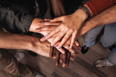 Çok ırklı bir arkadaş grubunun üst düzey bir görüntüsü. Takım çalışmasının, topluluk desteğinin ve dayanışmanın güçlü bir sembolü olarak ellerini üst üste koyuyorlar..