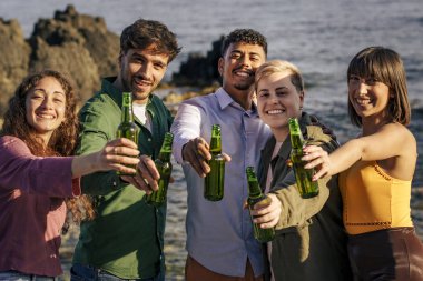 Beş genç arkadaştan oluşan mutlu çok ırklı bir grup, deniz kenarında yeşil bira şişeleriyle kutlama yaparken doğrudan kameraya bakıyorlar..
