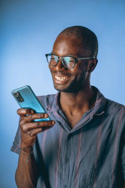 Gözlük takan neşeli bir adam akıllı telefonuyla meşgul oluyor, parlak bir şekilde gülümsüyor. Mavi arka plan neşeli atmosferi güçlendirir, bir bağ ve mutluluk anını yakalar..