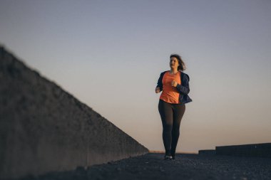 Gün batımında deniz kenarında gezinen genç bir kadının portresi spor giysisi ve kapüşonlu, aktif yaşam tarzı ve açık hava fitness konsepti giymiş..