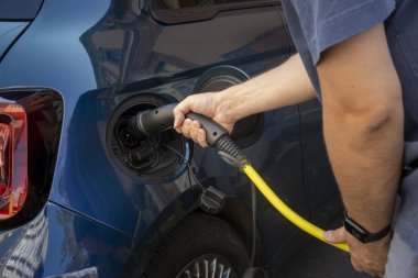 Şehir şarj istasyonunda sarı kabloyu mavi elektrikli arabaya bağlayan kişinin yakın çekimi, çevre dostu ulaşım.