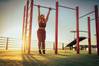 Jimnastik parkındaki yatay çubukta asılı duran sporcu kadının arka görüntüsü. Güneş ışığına karşı fotokopi alanı ile antrenman yapan bayan sporcu silueti..