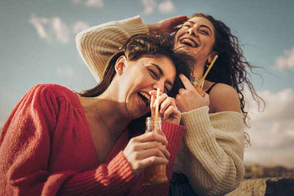 Two cheerful young women sharing a moment of genuine laughter while enjoying refreshing juice drinks outdoors on a bright day, capturing candid friendship and youthful happiness.