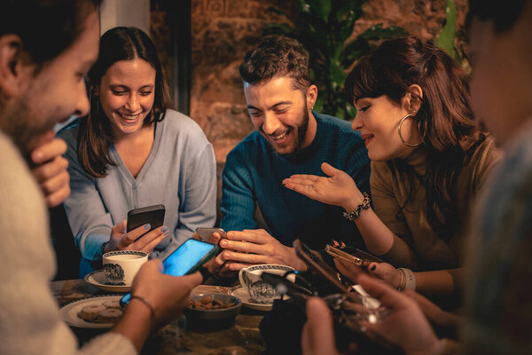 Happy group of young adults enjoying social time, sharing laughter and stories while looking at their mobile phones in an intimate cafe setting.