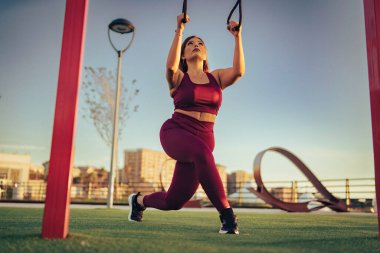 Askılarla süspansiyon egzersizi yapan genç bir kadının düşük açılı görüntüsü. Kadın fitness modeli gün batımında bacak egzersizi yapıyor..