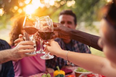 Gün batımında kadeh kaldırırken kırmızı şarap kadehlerini tokuşturan ellerin yakın görüntüsü. Sıcak arka plan ışığı, sığ alan derinliği, kutlama, arkadaşlık ve rahat sosyal atmosfer..