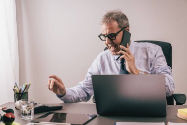 Üst düzey bir adam laptopuyla masasında çalışırken kızgın bir telefon görüşmesi yapıyor. İş stresi, iş baskısı, tükenme, çoklu görev ve zihinsel aşırı yüklenme kavramı.