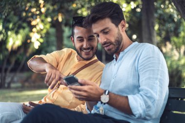 Parkta otururken akıllı telefona bakan iki erkek arkadaş. Paylaşılan dijital deneyim, teknoloji, bağlantı ve günlük arkadaşlık kavramı.