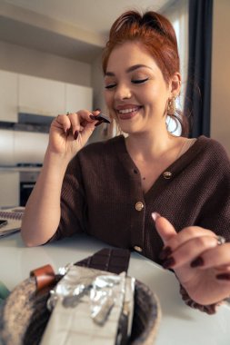 Modern bir mutfakta kırmızı saçlı, mutlu bir genç kadın bir parça siyah çikolatanın tadını çıkarırken gülümsüyor. Evdeki tatlı hoşgörü ve rahatlama anına odaklan..