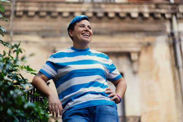 Joyful gender non-conforming individual with short blue hair and striped shirt laughing outdoors. Authentic expression of happiness and self-confidence against urban backdrop - diversity and inclusion