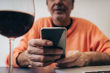 Turuncu kazaklı orta yaşlı bir adam, elinde bir dizüstü bilgisayar ve bir bardak kırmızı şarapla bir masada oturur ve modern teknoloji alışkanlıklarını ve dijital dengeyi göstermek için akıllı telefon ekranına odaklanır. ...