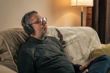 Olgun bir adam, kulaklıkları ve gözleri kapalı müzik dinlerken evdeki koltukta dinleniyor. Yavaş yaşam, zihinsel dinlenme, duygusal denge ve orta yaşamda sağlıklı yaşam tarzı kavramı.