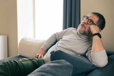 Evdeki kanepede dinlenen kıdemli bir adam düşünceli ve zihinsel olarak yorgun bir ifadeyle. Akıl sağlığı, stres, duygusal yorgunluk, iç gözlem ve sessiz iç mekan yaşam tarzı kavramı.