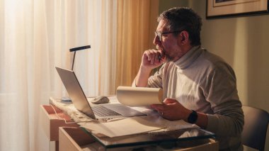 Evde çalışan kıdemli bir profesyonel, bilgisayar kullanırken basılı belgeleri inceliyor. Uzaktan çalışma ortamında deneyim, karar verme, liderlik, stratejik düşünme ve profesyonel sorumluluk kavramı.