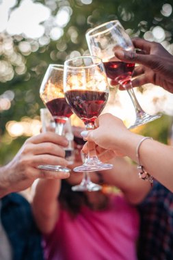 Gün batımında çeşitli ellerin kırmızı şarap bardaklarını tıkırdatmasıyla. Sıcak yaz ışığı, bokeh arka plan ve yaşam tarzı kutlama konsepti.