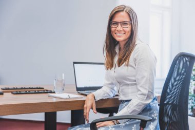 Ofiste evrak işleriyle uğraşırken bilgisayarını ve bilgisayarını kullanarak başarılı bir şekilde gülümseyen iş kadını.