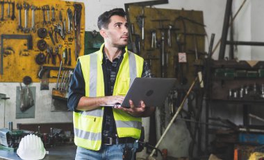 Üniformalı ve miğferli genç bir mühendis, bilgiye bakmak ve metal fabrikasındaki torna makinelerine bakmak için dizüstü bilgisayar tutuyor..