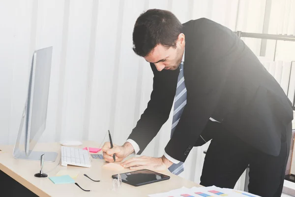 A businessman wrote a post-it on a wooden desk with a computer on the front, a tablet and a credit card next to it. The business man\'s hands writing a post-it to list and plan an event in the office.