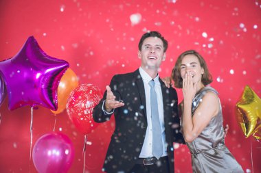 Önündeki konfetiler arasında genç çift mutlu. Yanlarında renkli balonlar ve yılbaşı ve yılbaşı konseptinde kırmızı arka plan var..