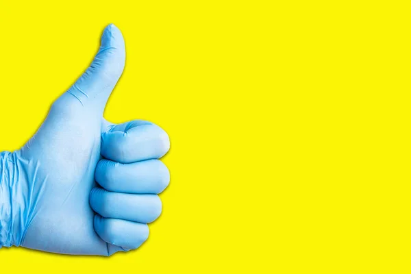 Thumb up approval symbol. Hand in blue latex glove on yellow background showing OK sign. Empty copy space medical background. Doctor successful operation. Virus epidemic hygiene glove.