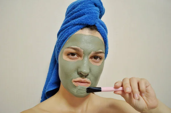 A woman with a brush applies a clay mask to her face with a blue towel on her head