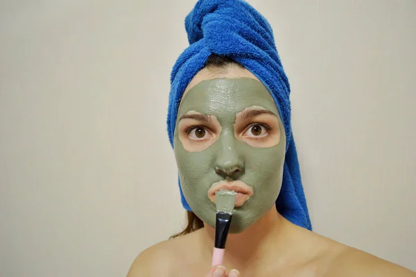 A woman with a brush applies a clay mask to her face with a blue towel on her head