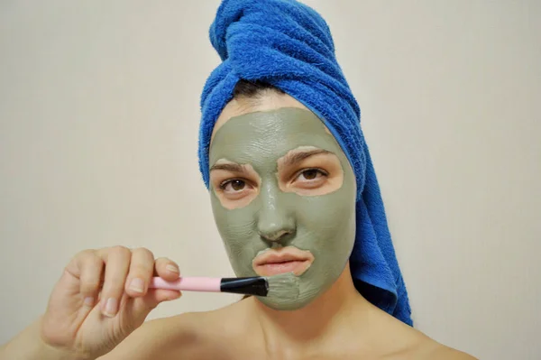 A woman with a brush applies a clay mask to her face with a blue towel on her head