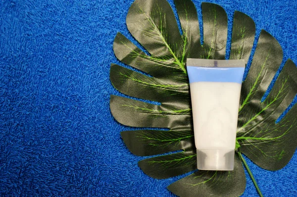 White tube with cream lies on a palm leaf against a background of blue fabric.
