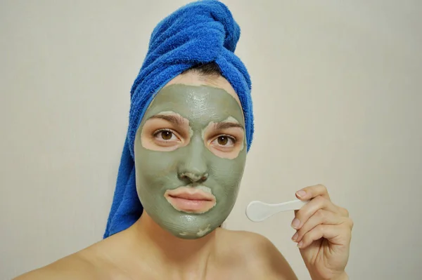 Woman with a spatula applies a clay mask to her face with a blue towel on her head