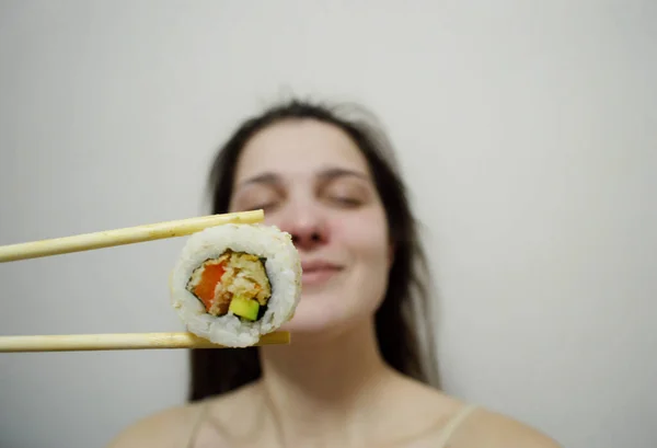 Beautiful young woman eating sushi with wooden chopsticks on a light background.