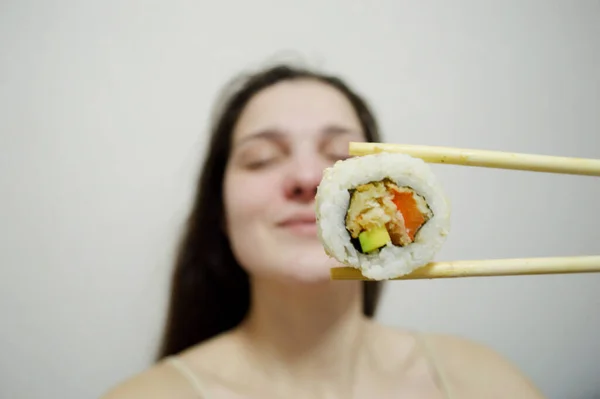 Beautiful young woman eating sushi with wooden chopsticks on a light background.