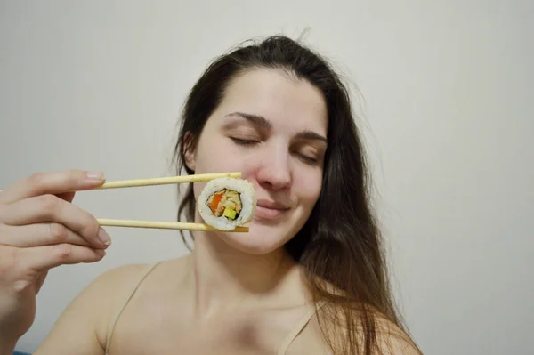 Beautiful young woman eating sushi with wooden chopsticks on a light background.