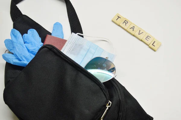Black belt bag with passport, medical mask, blue rubber gloves and sunglasses on white background. Travel word made from wooden cubes
