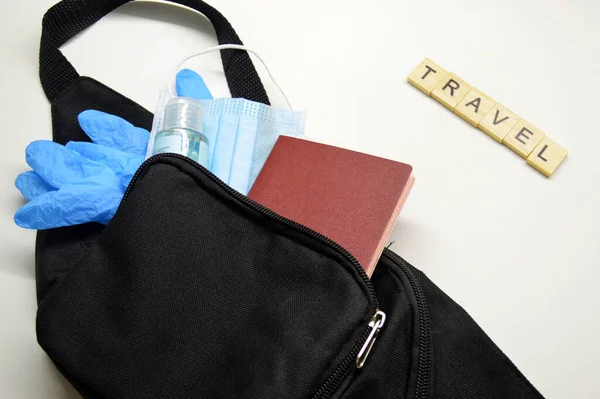 Black belt bag with passport, medical mask, blue rubber gloves and antiseptic on white background. Travel word made from wooden cubes