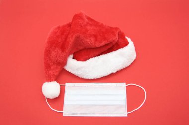 Noel için yaratıcı bir tıbbi kart. Kırmızı arka planda Noel Baba şapkası ve tıbbi maske. Yeni yılda tıp kavramı. Yukarıdan manzara. Dümdüz. Kopyalama alanı.