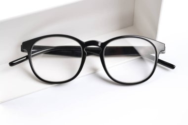 Siyah çerçeveli optik gözlükler beyaz arka planda beyaz bir hediye kutusunun yanında yer almaktadır. İndirim, promosyon, optik mağazalarda satış, gözlük onarımı, gözlük hediye olarak gözlük.