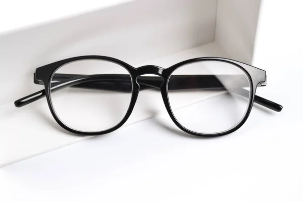 Siyah çerçeveli optik gözlükler beyaz arka planda beyaz bir hediye kutusunun yanında yer almaktadır. İndirim, promosyon, optik mağazalarda satış, gözlük onarımı, gözlük hediye olarak gözlük.