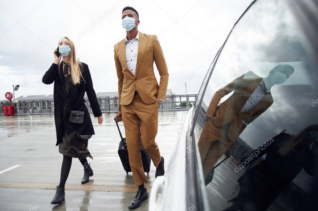 Business Couple Wearing Masks Outside Airport For Business Trip With Luggage During Health Pandemic