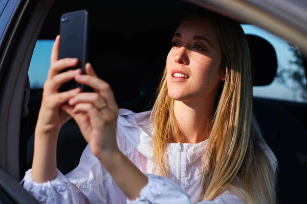 Young woman on road trip vacation taking photo out of rental car window on mobile phone