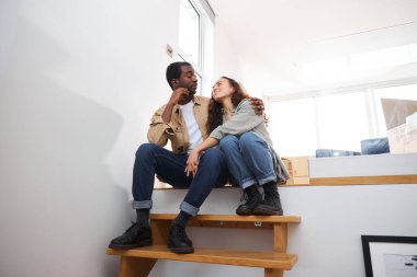 Genç, etnik kökenli bir çift yeni evin taşınma gününü kutluyor. Merdivenlerde oturuyor. Birlikte kaldırma kutularıyla çevrili.