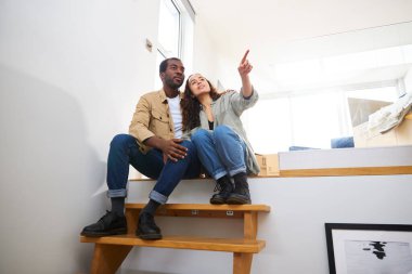 Genç, etnik kökenli bir çift yeni evin taşınma gününü kutluyor. Merdivenlerde oturuyor. Birlikte kaldırma kutularıyla çevrili.