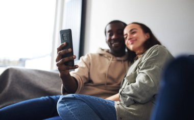 Genç etnik kökenli çift evdeki kanepede dinleniyor cep telefonunda selfie için poz veriyor.