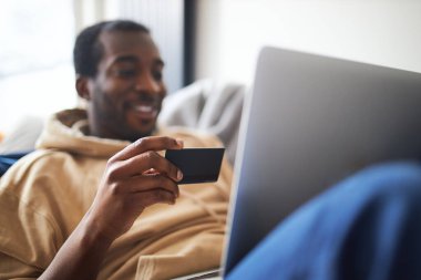 Evde dizüstü bilgisayarla kanepede uzanmış, ön plana odaklı internet alışverişi yapmak için kredi kartı kullanan melez bir gencin yan görüntüsü