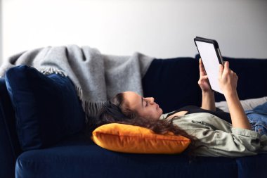 Dijital tablet üzerine kitap okuyarak kanepede uzanan gülümseyen etnik kökenli genç kadının profil görüşü