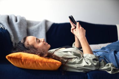 Sosyal medyayı cep telefonundan kontrol etmek için evdeki kanepede uzanmış gülümseyen melez kadının profil görüşü