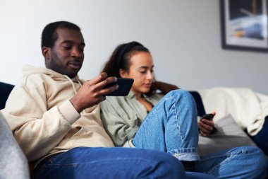 Genç, etnik kökenli bir çift evdeki kanepede oturuyor. Sosyal medyayı cep telefonlarıyla kontrol ediyor ama birbirlerini görmezden geliyorlar.